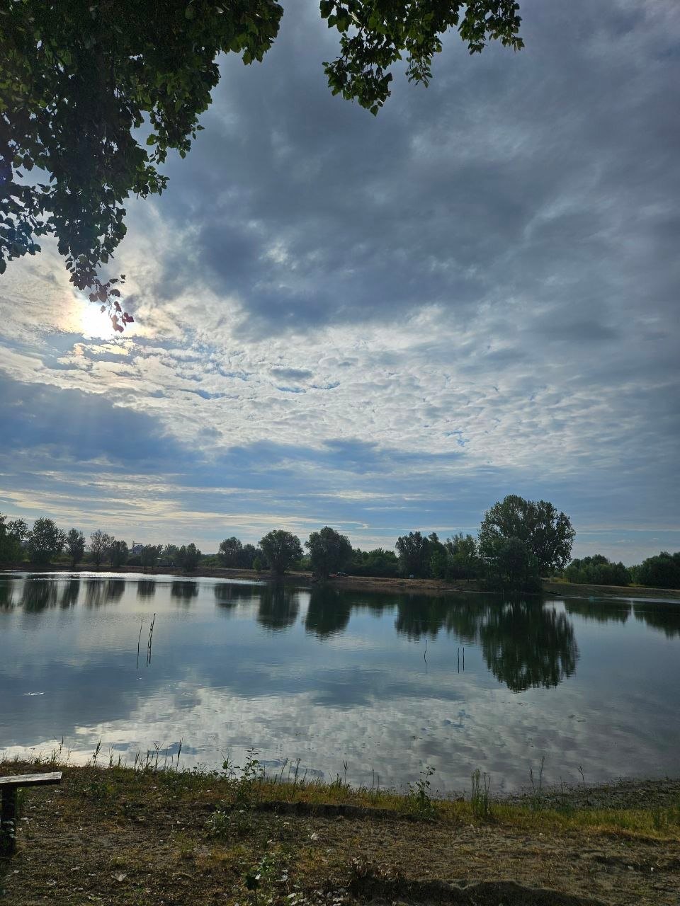 The sun behind clouds over Lake Provala, the sky and trees mirrored in the lake